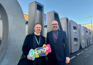 Two people standing in front of a metal sign that says 'Edinburgh' and one on the left holding a Change Mental Health sign.