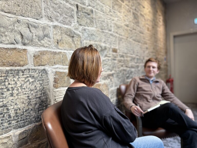 Two people in a stone-walled hallway; foreground person with short brown hair seen from behind, another person seated with a notebook in the background.