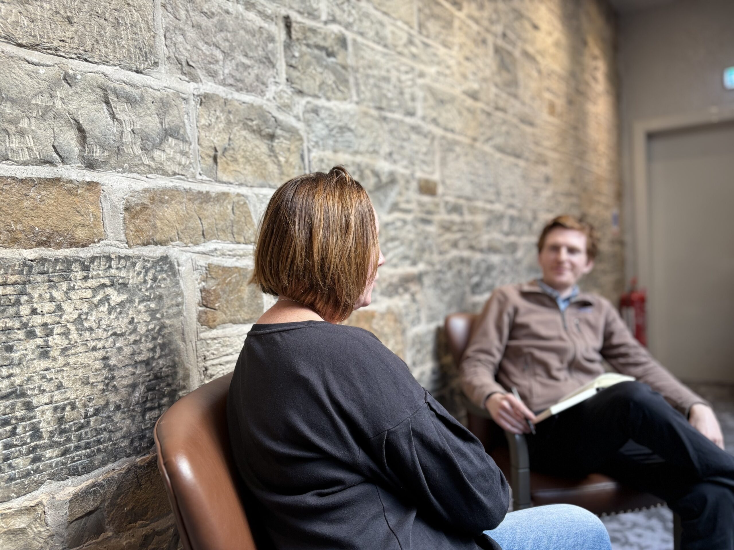 Two people in a stone-walled hallway; foreground person with short brown hair seen from behind, another person seated with a notebook in the background.