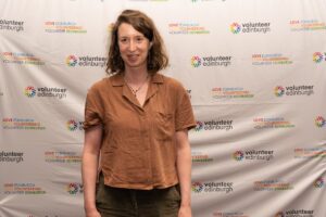 Smiling woman in a brown button-up shirt poses in front of a Volunteer Edinburgh backdrop. Rosa, creative writing volunteer