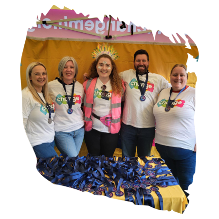 Five smiling people posing together at a booth, wearing white shirts with a colorful 'change' logo; ribbons and badges spread on a yellow table in front of a pink banner and backdrop.