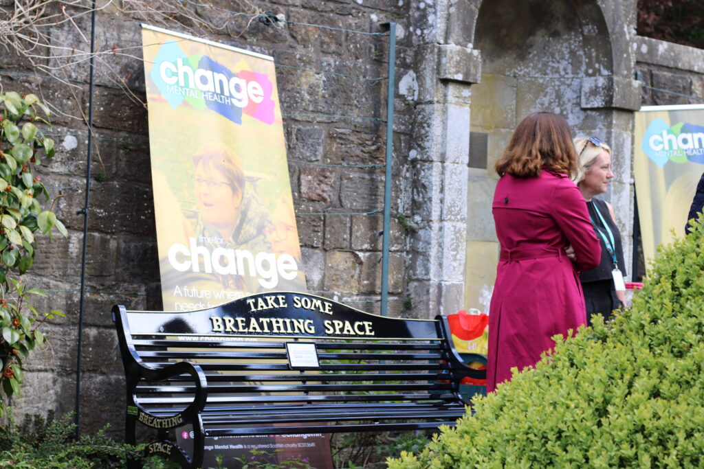 Outdoor scene with Change Mental Health banners on a stone wall and two women talking beside a black curved bench.