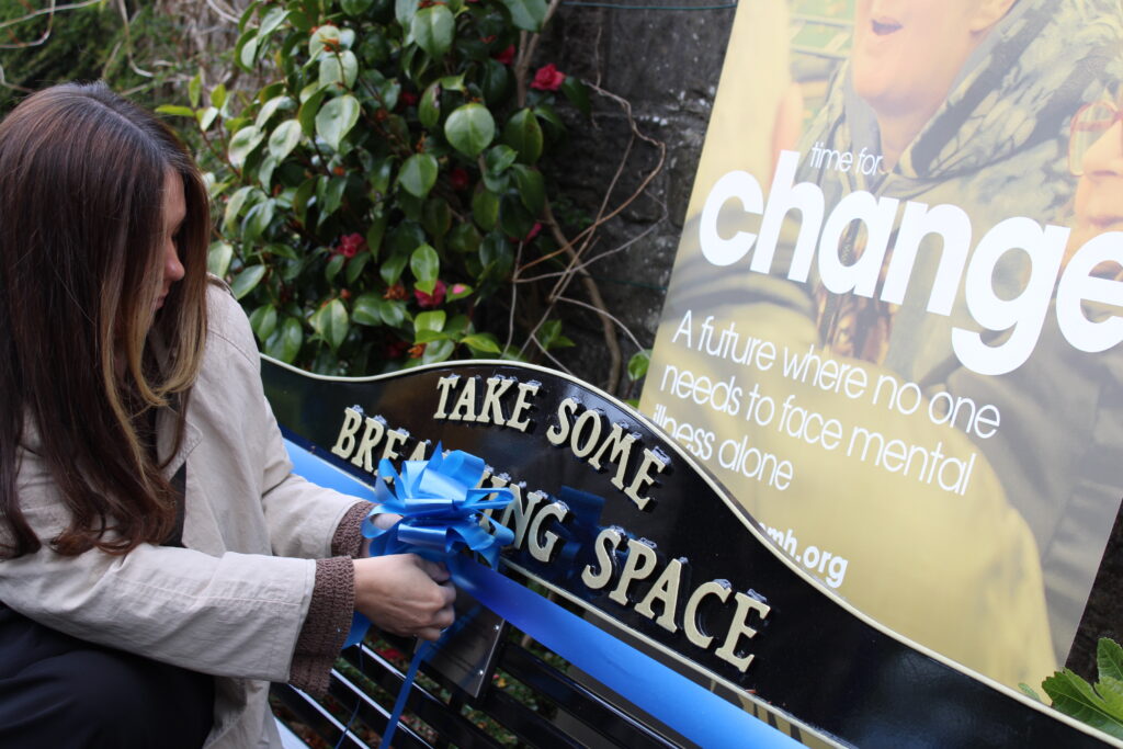 Person with long brown hair ( Natalie, NHS24 Breathing Space) ties blue ribbons onto a black-and-yellow banner outdoors, promoting change in the campaign.