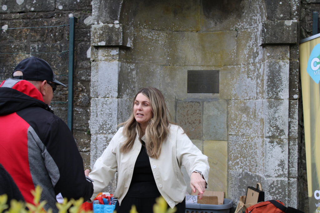 Woman in a beige jacket (Susan Grant, Change Mental Health) talks to a man in a red/black jacket at an outdoor charity stall with boxes and baskets against a mossy stone wall behind them.
