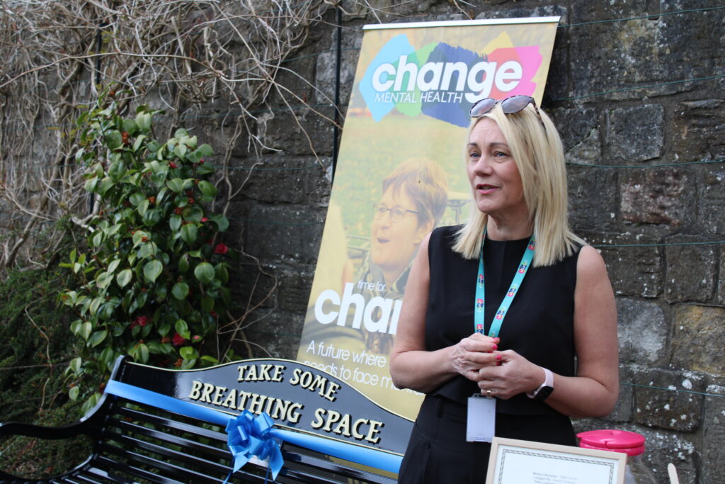 Blonde woman in a black outfit (Lynne Penman) with a lanyard stands beside a colorful Change Mental Health banner outdoors by a stone wall.