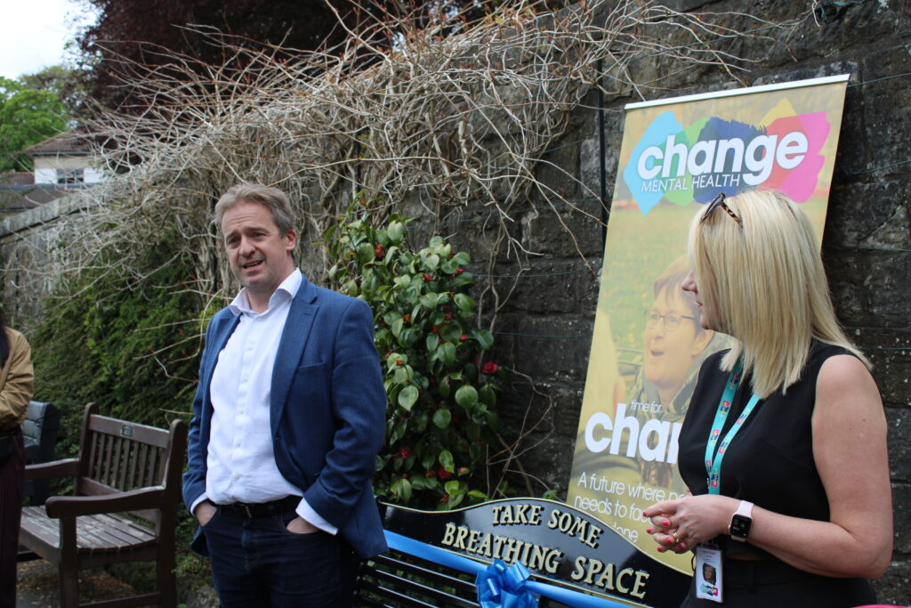 Man in a blue blazer (Greame Downie MP) and a blonde woman with a lanyard ( Lynne Penman) stand beside a colorful Change Mental Health banner outdoors.