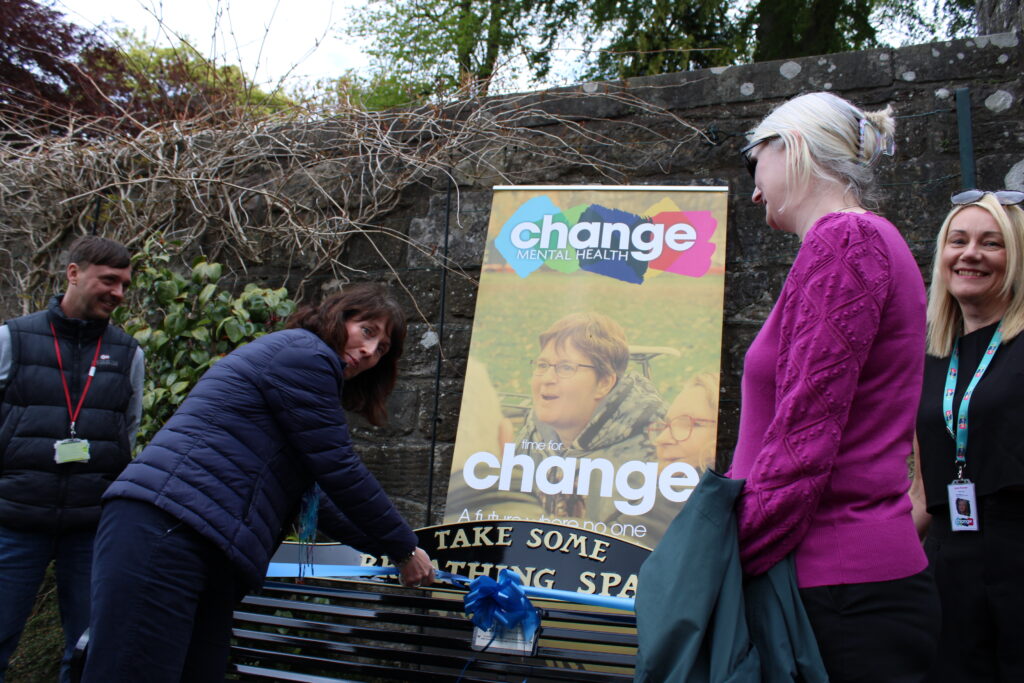 Adults gather around a colorful Change Mental Health poster outside, standing near a bench and stone wall. From left: representative of Fife Council, two representatives of Carnagie Dunfermline Trust and Lynne Penman from Change Mental Health