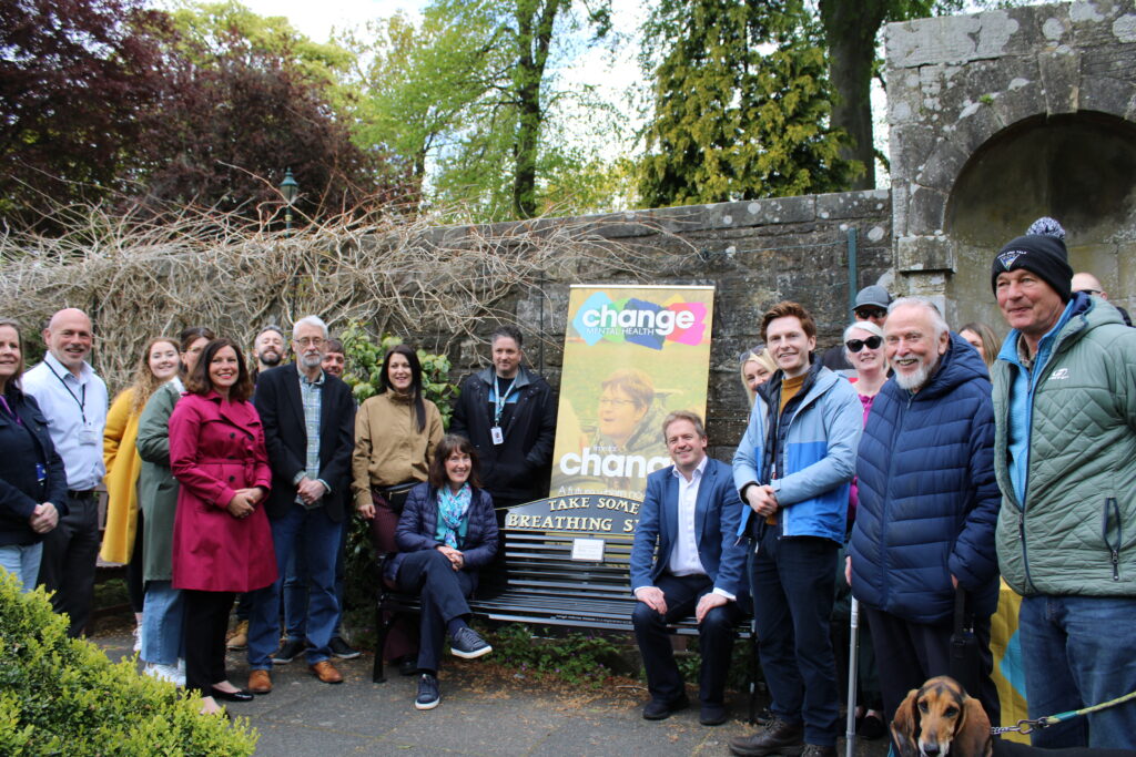 Group of diverse adults posing for a photo outdoors beside a colorful 'change' mental health banner on a Take Some Breathing Space bench in a garden setting.
