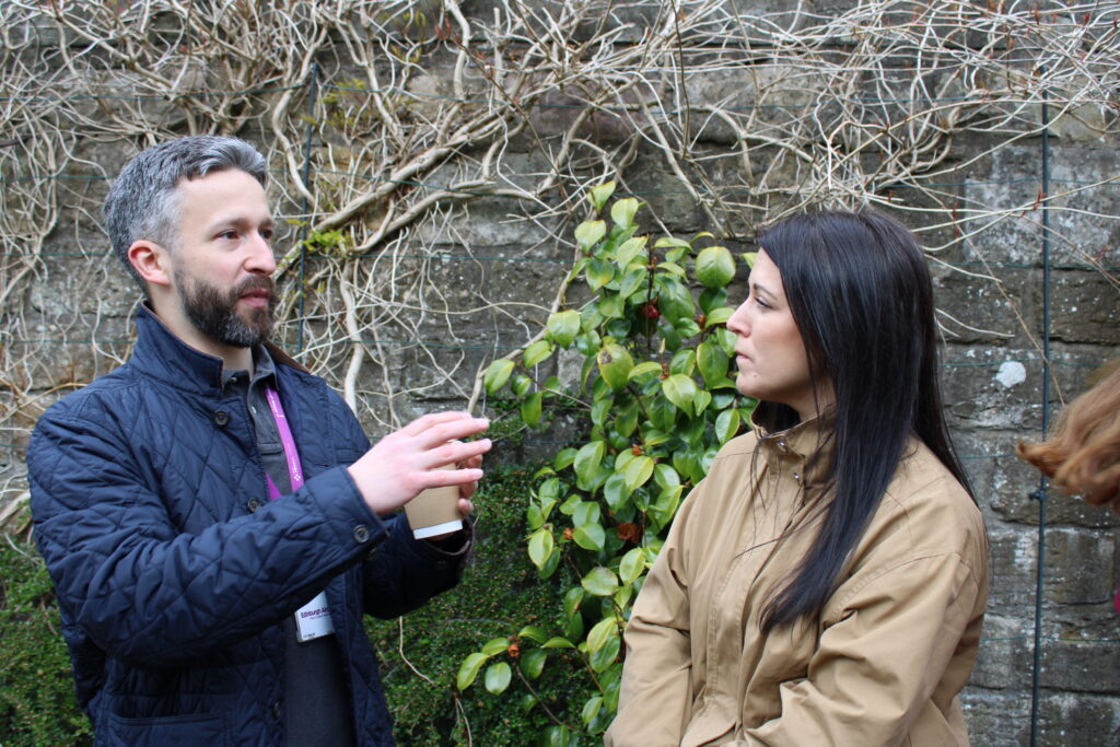 Man with short gray hair (Aaron- Edinburgh Airport) holds a coffee cup while talking to a woman (Lisa-Jane Dock, NHS24 Breathing Space) outdoors in front of a stone wall and vines.
