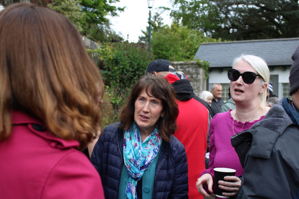 Group of adults (Carnegie Dunfermline Trust representatives) chatting at an outdoor gathering; woman in pink top with sunglasses holds a disposable cup on the right side.