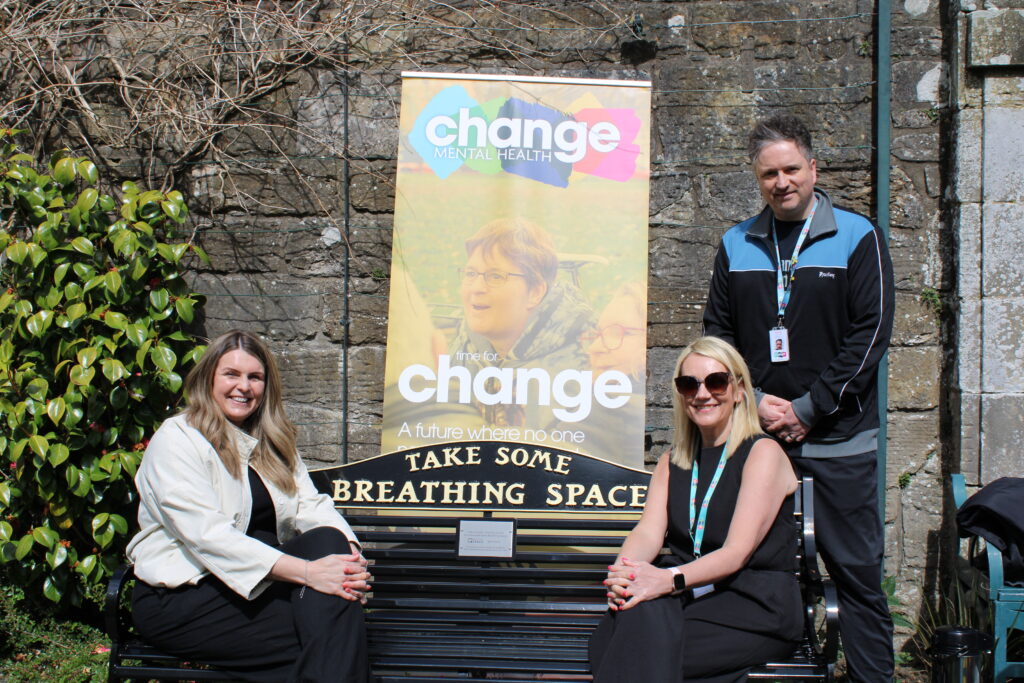 Three people (from left Susan Grant, Lynne Penman and standing Barry Kydd) sit on a bench beside a colorful 'change mental health' banner outdoors against a stone wall.