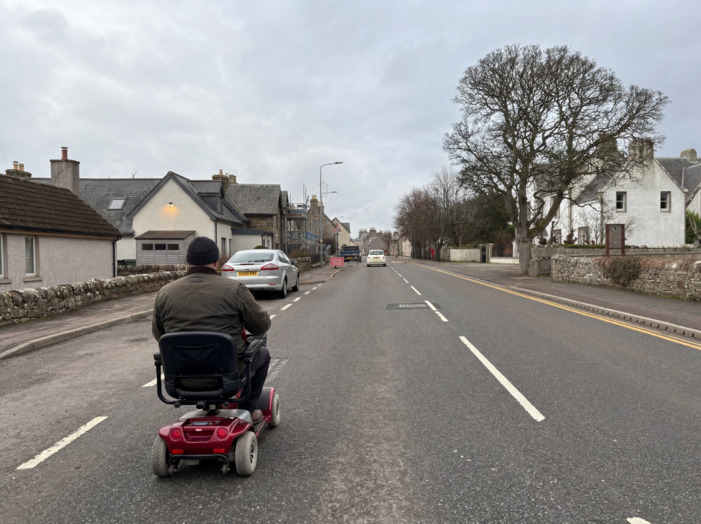 Men on the mobility scooter in dark jacket and hat on the road in the a village - Thomas’s story: Long-term caring and access to local, community support