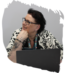 Smiling woman with short dark hair and glasses rests her chin on her hand while seated at a laptop with a lanyard around her neck.