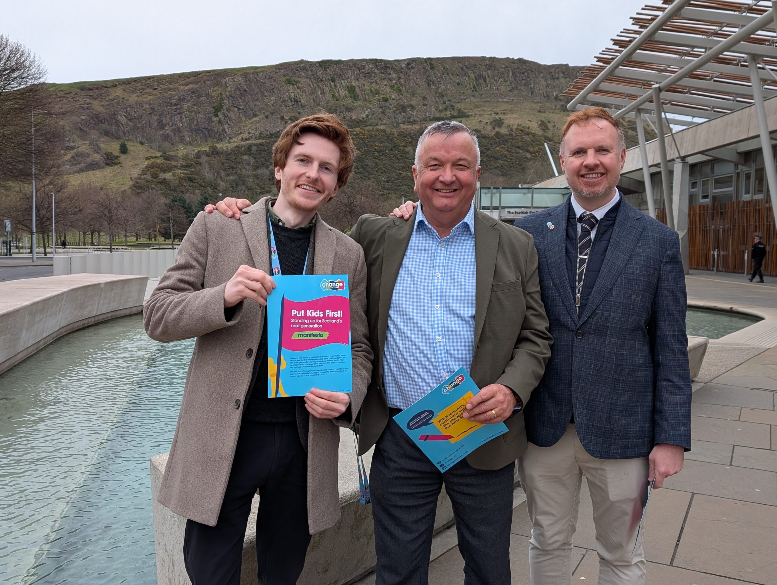 Three men in smart casual jackets stand together outdoors, holding bright Change Mental Health manifesto in front of Scottish Government building - symbolising bringing mental health policy into action in Scotland