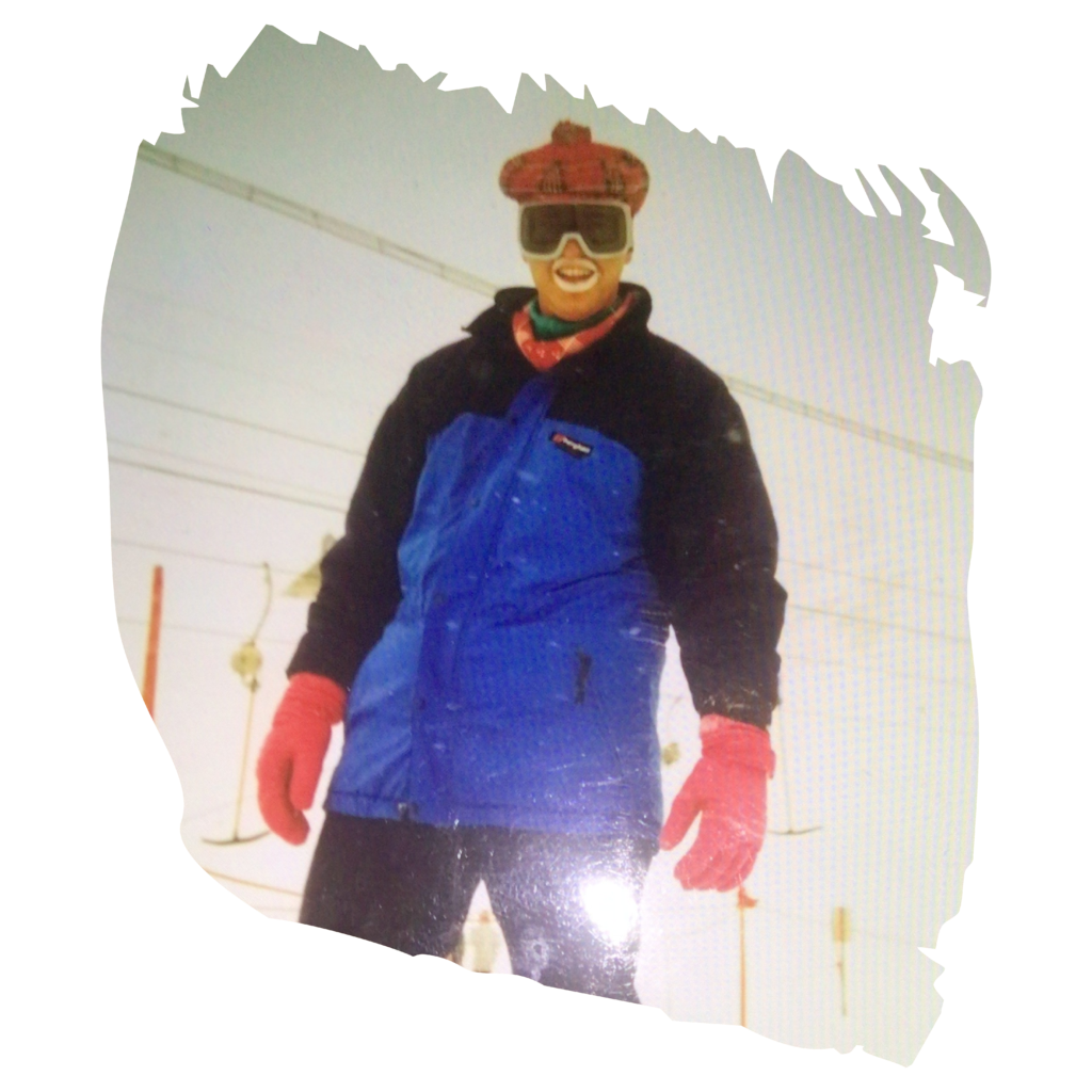 Person in blue ski suit and red gloves standing on a snowy slope, wearing goggles and smiling at the camera.