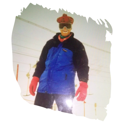 Person in blue ski suit and red gloves standing on a snowy slope, wearing goggles and smiling at the camera.