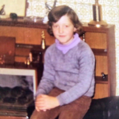 Young girl sits on the floor in a retro living room, wearing a purple turtleneck and gray sweater.