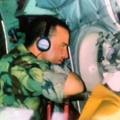 Soldier in camouflage uniform wearing a headset, seated inside an aircraft cockpit beside control panels.