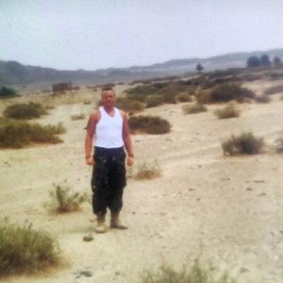 Man standing in a sandy desert with sparse shrubs, wearing a white sleeveless shirt and dark cargo pants.