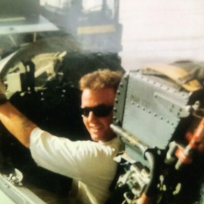 Man wearing sunglasses sits in the cockpit of a military aircraft, smiling at the camera with one arm resting on the side panel.