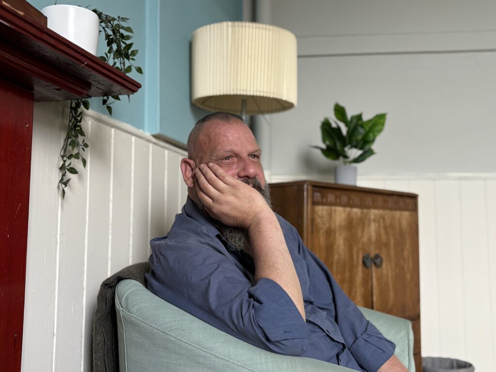 Smiling, bearded man with hand on his cheek sits in a teal-walled room, wearing a blue shirt.