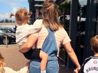 A woman in denim overalls carries a young child while two other kids walk beside her near a storefront ATM.