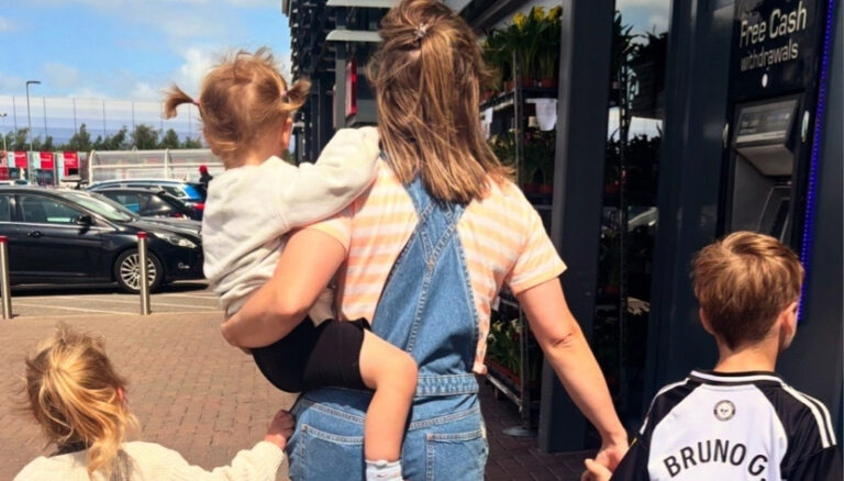 A woman in denim overalls carries a young child while two other kids walk beside her near a storefront ATM.