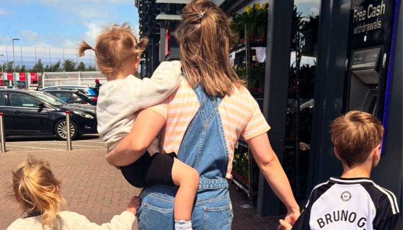 A woman in denim overalls carries a young child while two other kids walk beside her near a storefront ATM.