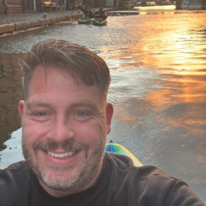 Jonny portrait Smiling man taking a selfie on a boat canal at sunset with water reflecting orange light behind him.