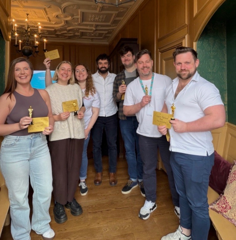 Group of seven smiling adults posing in a wood-paneled room, each holding small gold award plaques and certificates.