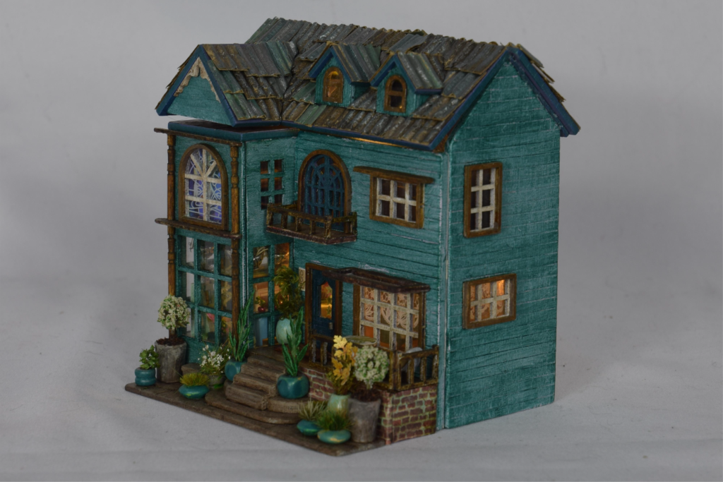 Teal two-story miniature house with slate roof and a small front garden with steps and potted plants.