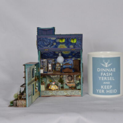A detailed miniature multi-story dollhouse with tiny rooms and furniture, placed beside a white mug with a blue label.