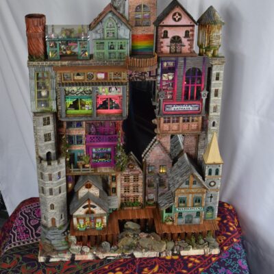 Colorful multistory miniature town made of stacked houses with storefronts, towers, and a rocky base on a patterned cloth.