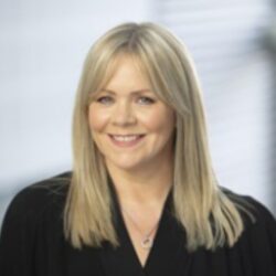 Carolann Begbie - Professional headshot of a smiling blonde woman wearing a black top, with a softly blurred office background.