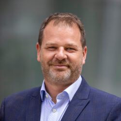 Chris Cummings - Professional headshot of a middle-aged man in a navy blazer and light blue shirt, smiling outdoors.
