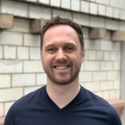 Euan Wielewski -Smiling man with short dark hair and a beard, wearing a navy shirt, in front of a light brick wall.