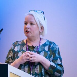 Fiona Hogg - Woman with short blonde hair wearing glasses on her head speaks at a podium during a conference, wearing a teal geometric-patterned dress.
