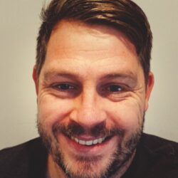 Jonny Cobbold -Smiling man with short dark hair and beard looking at the camera, neutral background.