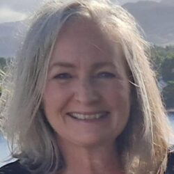 Judith Mackinnon- Smiling woman with shoulder-length gray hair, outdoors with trees behind her.