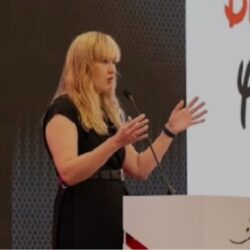 Marzena D’Arcy - Woman with long blonde hair wearing a black dress speaks at a podium with a microphone on stage, gesturing with her hands during a presentation.