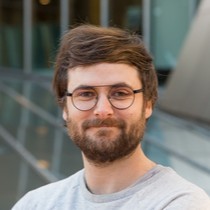 Milo Phillips-Brown - Portrait of a smiling man with glasses and a beard, wearing a light gray shirt, in front of a glass-walled building.