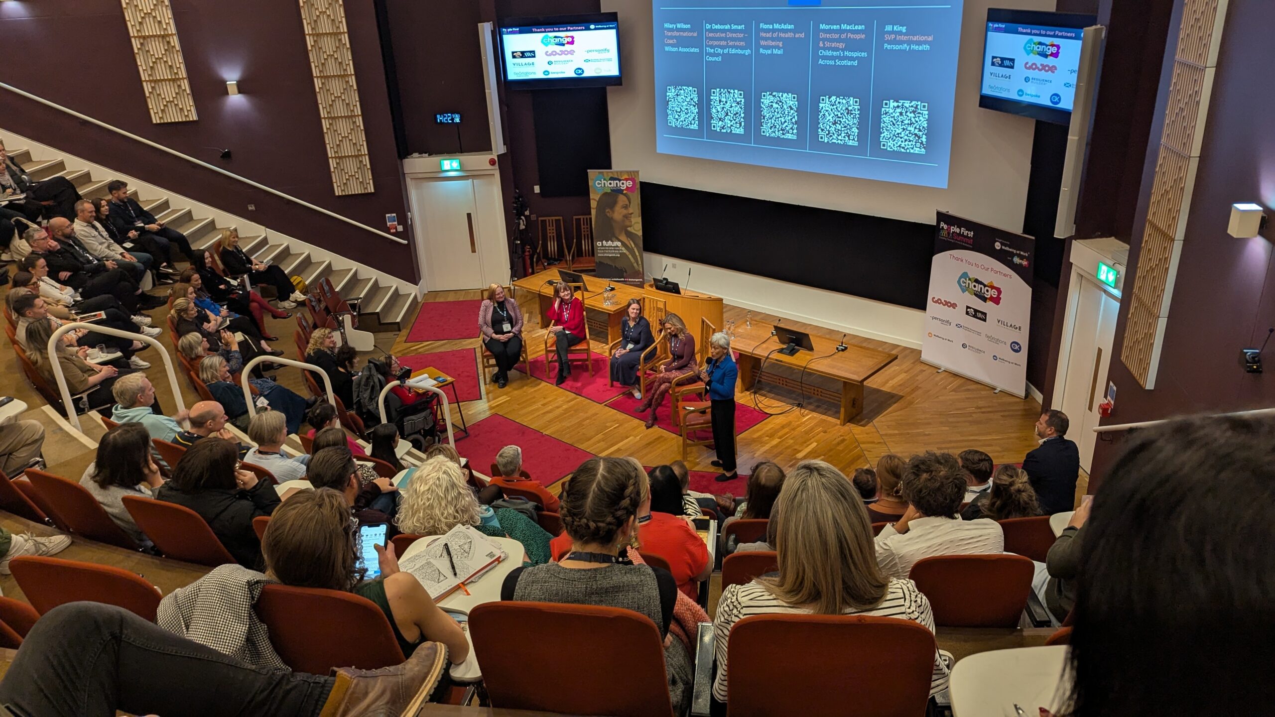 Panel discussion on stage with five seated panelists and a presenter, in a lecture-hall style auditorium; large screen behind shows slides.