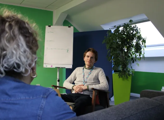 Man wearing a lanyard sits in an office chair for a casual meeting, with a whiteboard, green and blue walls, and a tall plant in a lime pot behind him.