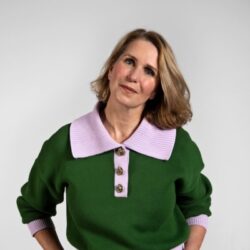 Vicky Hope - Portrait of a woman with blonde hair wearing a green sweater with a pink collar, posing against a light gray background.