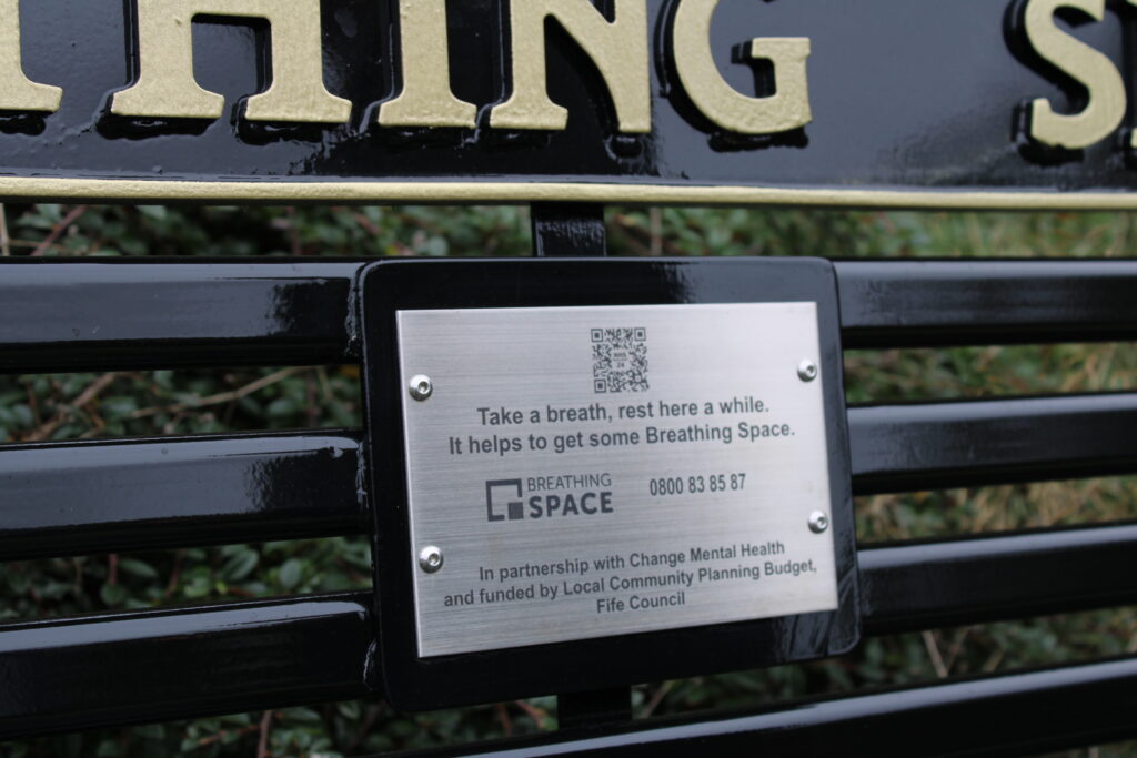 Close-up of a metal information plaque on a black bench, featuring a QR code and the text about Take a breath, rest here a while (Breathing Space).