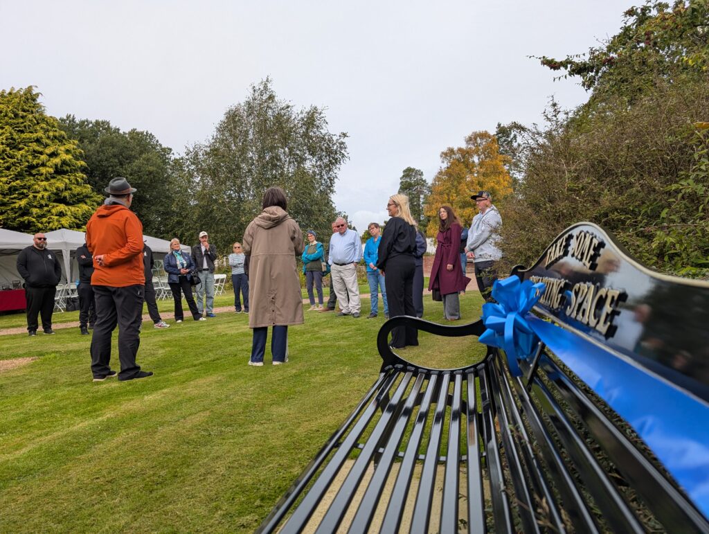 A group of people standing on a grassy lawn at an outdoor event, with trees behind and a blue and black bike rack on the right foreground.