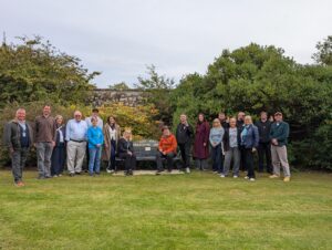 Take Some Breathing Space bench opening by Change Mental Health in St. Andrews - Group of about 20 people standing in a grassy park, posing for a photo in front of a stone wall and dense shrubs behind them.