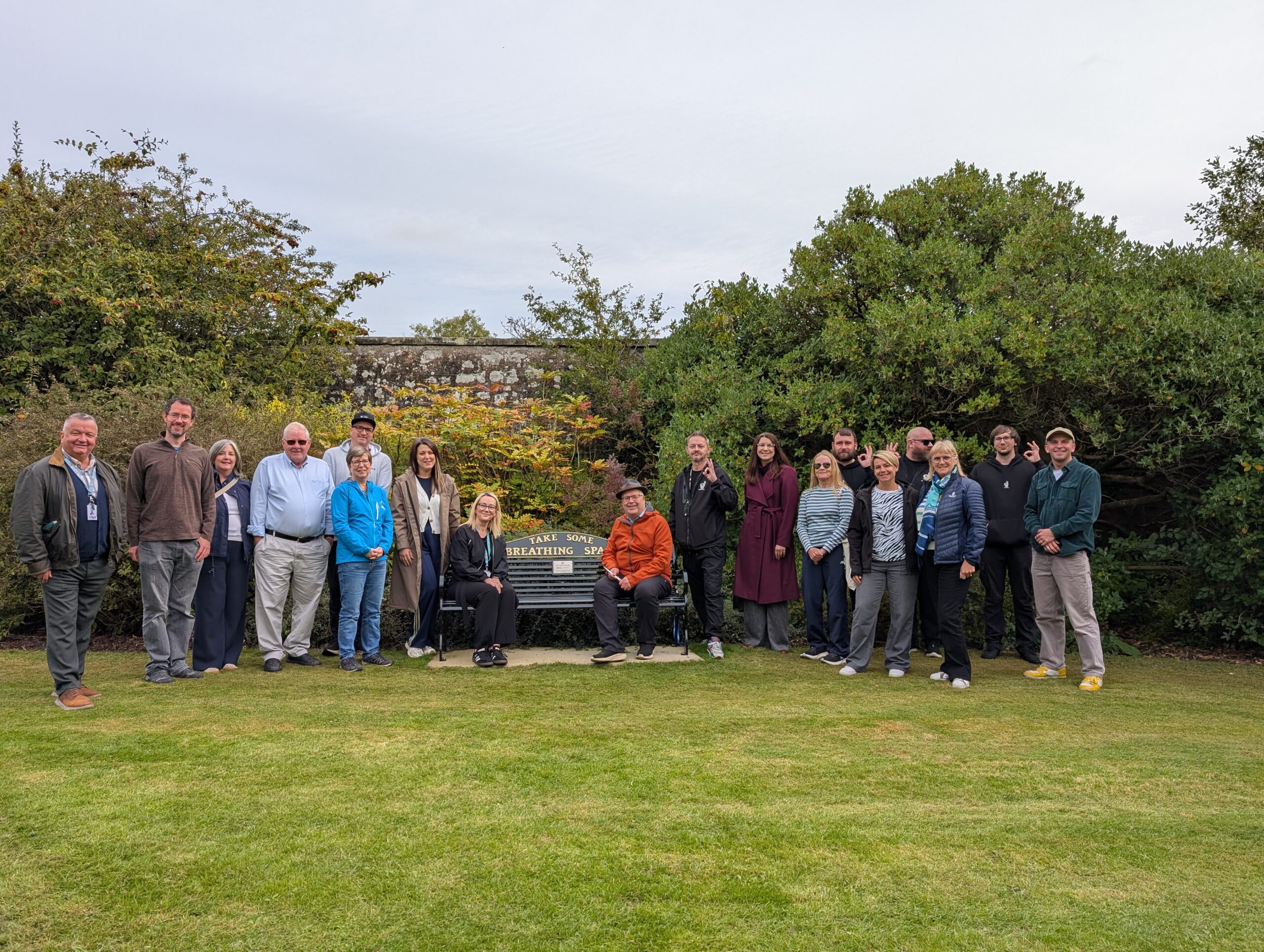 Take Some Breathing Space bench opening by Change Mental Health in St. Andrews - Group of about 20 people standing in a grassy park, posing for a photo in front of a stone wall and dense shrubs behind them.