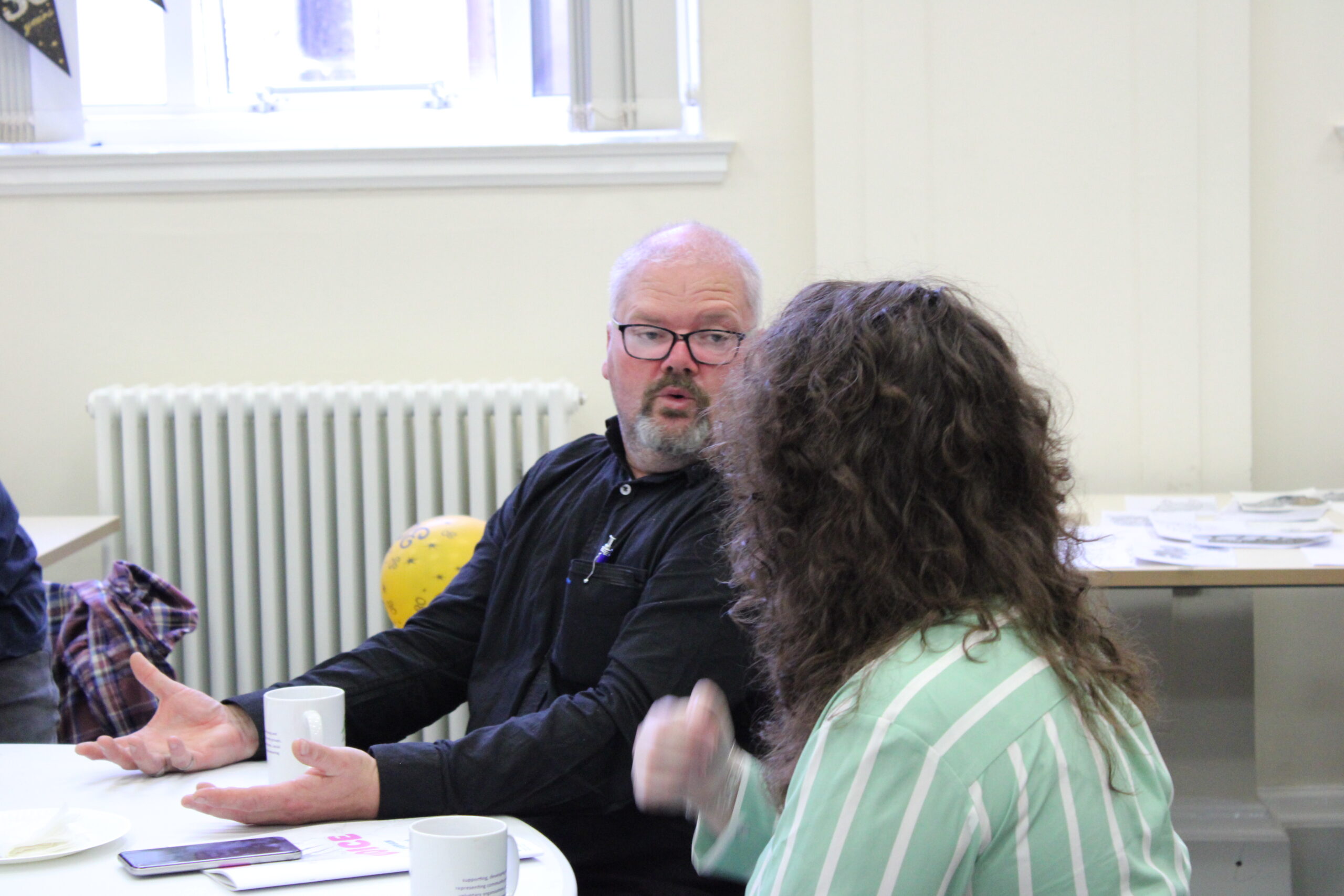 Man with glasses wearing a black shirt speaks at a table, hands gesturing as others listen in a bright office.