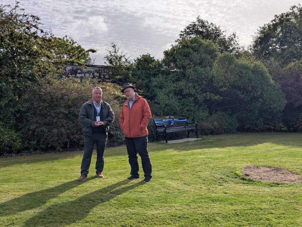 Two men standing on a grassy lawn in a park, with dense shrubs and a bench behind them.